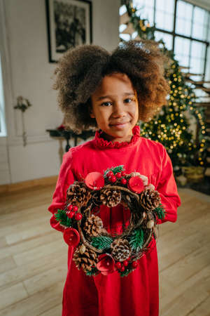 Little girl with curly hair holding decorative wreath made from dry branches and cones. Idea for DIY handmade festive decoration. Selective focus.の写真素材