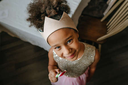 Girl wearing toy crown eating Christmas candy cane standing near the table at home.の写真素材