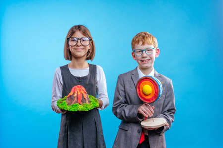 Two schoolchildren with eye glasses holding their scientific projects volcano and section of the Earth on blue background with blank space for textの写真素材