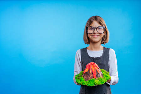 Schoolgirl holding her scientific project volcano on blue background with blank space for textの写真素材