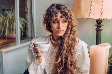 Portrait of young woman wearing vintage blouse sitting at the cafe drinking tea or coffee. Selective focus.の写真素材
