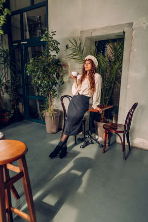 Young woman with curly hair wearing vintage style blouse with collar and beret at the cafe drinking tea or coffee.の写真素材