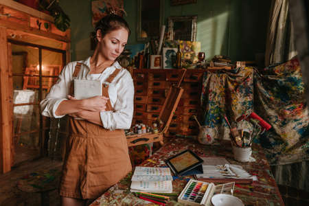 Lifestyle portrait of female artist standing near worktable holding her sketchbookの写真素材