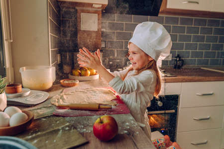 Little girl wearing apron and chef's hat rolling the dough with a rolling pin playing with flour on the table while preparing the apple pie in the kitchen. Selective focus.の写真素材
