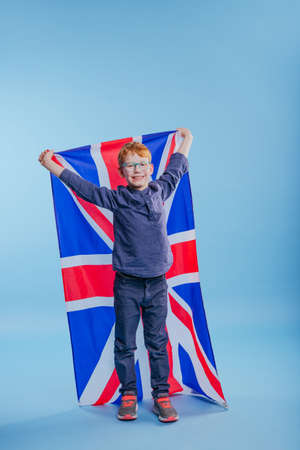 Schoolboy wearing eye glasses holding Great Britain Union Jack flag on blue background with blank space for textの写真素材