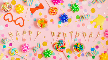 Multicolored lollypops, confetti, bows and decorative paper crown on stick for photosession on pink background with gold candles letters Happy birthday with blank space for text. Horizontal panoramic banner. Top view, flat lay.の写真素材
