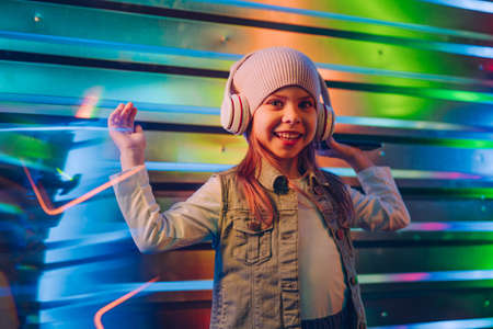 Little girl wearing headphones using smartphone standing on the colorful neon lights background. Generation Alpha using technology. Selective focus on the face.の写真素材