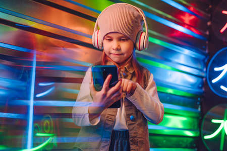 Little girl wearing headphones using smartphone standing on the colorful neon lights background....