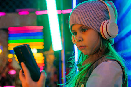 Little girl wearing headphones using smartphone standing on the colorful neon lights background....