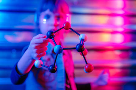Little girl holding molecular model standing on the colorful neon lights background. Generation Alpha using technology. Selective focus on the plastic balls.の写真素材