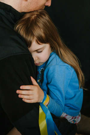 Portrait of frightened little girl with Ukrainian flag ribbon on her hand hugging her father. Peace and pray for Ukraine's children. Selective focus on the girl's face.の写真素材