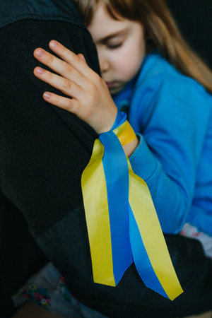 Portrait of frightened little girl with Ukrainian flag ribbon on her hand hugging her father. Peace and pray for Ukraine's children. Selective focus on the ribbon.の写真素材