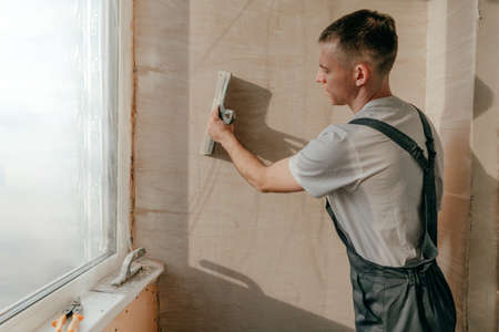 Young man builder dressed in work clothes overall applying putty mortar on the balcony wall with a spatula.の写真素材