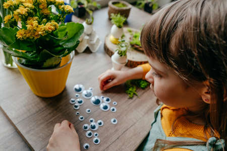 Little girl decorating eggshell with toy eyes on the wooden table at home. Creative DIY idea for festive Easter decoration. selective focus.の写真素材