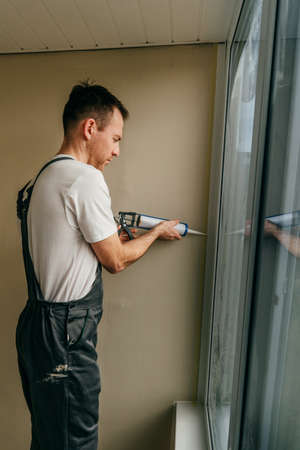 Young man wearing overalls sealing cracks between window and trim using waterproof silicone caulk on...