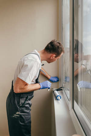 Young man wearing overalls sealing cracks using waterproof silicone caulk on the balcony. Removing...