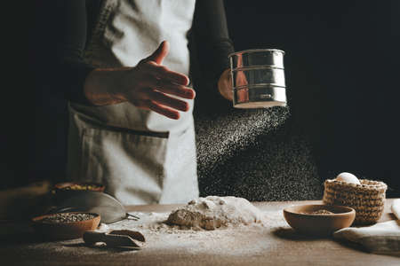 Unrecognizable young man sifting flour for kneading dough. Males hands making bread on dark background. Selective focus on the hands.の写真素材
