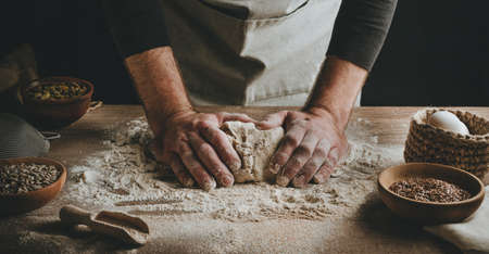 Unrecognizable young man kneading dough on wooden table. Males hands making bread on dark background. selective focus.の写真素材