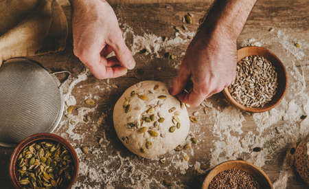 Unrecognizable young man making whole wheat bread sprinkling sunflower and pumpkin seeds on wooden...