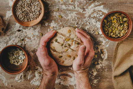 Unrecognizable young man making whole wheat bread with sunflower and pumpkin seeds on wooden background. selective focus.の写真素材