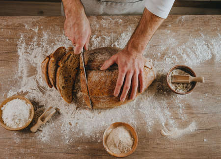 Unrecognizable young man in apron cutting homemade whole grain bread. selective focus. Top view,...