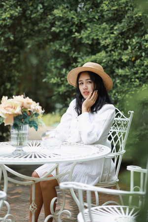 Young woman sitting in a restaurant, wearing a hat and white dressの写真素材