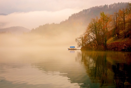 A boat and island in middle of lake Bled with colorful trees in autumn on foggy day