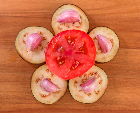 Slices of eggplant with cloves of garlic and a half of tomato on a wooden board surface.の写真素材