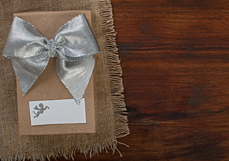 Wrapped gift box with silver ribbon bow on a dark walnut wood, on linen napkin with white card and silhouette of angel, top view, close upの写真素材