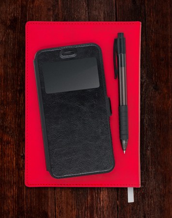 Composition with smartphone and pen on a red diary notebook placed on a dark wooden surfaceの写真素材