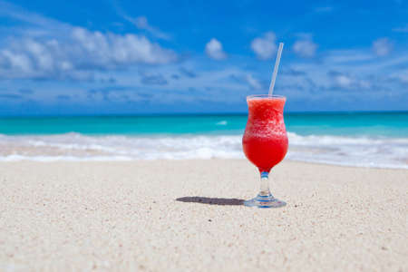 red drink with a straw on the white beach with ocean and sky in the background