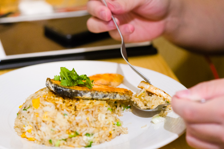 fried sliced salmon with fried rice.man hands use fork and spoon for fusion food.sliced salmon with rice on white plate in restaurant or cafe.man sitting and eating sliced salmon on laptop backgroundの写真素材