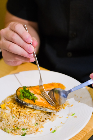 fried sliced salmon with fried rice.man hands use fork and spoon for fusion food.sliced salmon with rice on white plate in restaurant or cafe.man sitting and eating sliced salmon on laptop backgroundの写真素材