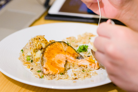 fried sliced salmon with fried rice.man hands use fork and spoon for fusion food.sliced salmon with rice on white plate in restaurant or cafe.man sitting and eating sliced salmon on laptop backgroundの写真素材