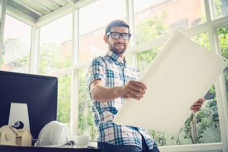 Young caucasian holding his blueprint project at the desk in home office.