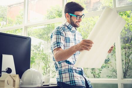 Young caucasian holding his blueprint project at the desk in home office.の写真素材