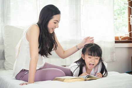 Happy Asian mother and daughter having quality time by reading book together on bed.の写真素材