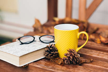 Close up photo of glasses, yellow mug with tea, pine cone, book and orange fallen leaves on wooden table outside. Comfort, coziness and hygge concept. Autumn mood. Still life. Selective focusの写真素材