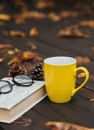 Yellow cup of tea with a book and glasses on a wooden background with a lot of dry fallen leaves. Relaxing scene. Selective focus. Bad weather.の写真素材