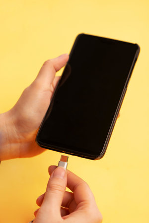 Hand connecting type c charging cable to the smartphone on yellow background close up.の写真素材