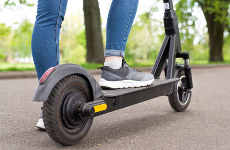 Close up woman legs standing on the electric kick scooter in summer day back view riding.
