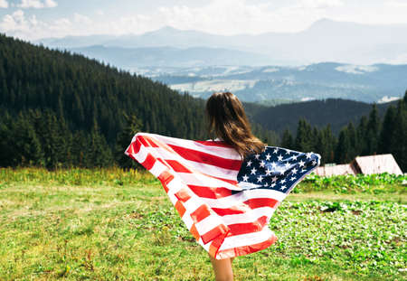 Back view young woman with american flag in hands standing on top of mountain. Female hiker waving national usa flag outdoor. Patriotic girl celebrating us independence day at vacation.の写真素材
