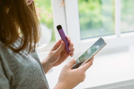 girl holds and smokes a purple disposable electronic cigarette by the window. Modern communication...