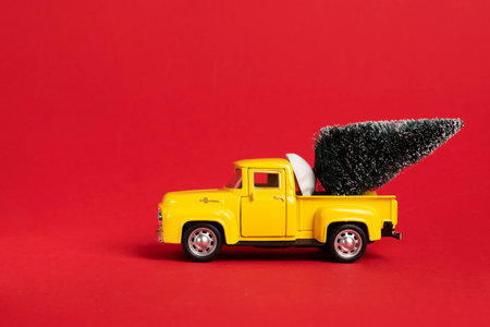 Yellow retro toy pickup carrying a Christmas tree on red background. Christmas and New Year celebration concept. Copy space, selective focusの写真素材