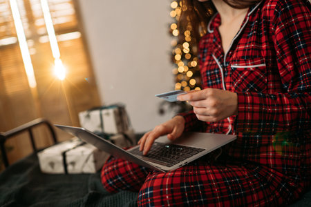 Woman shopping online in festive pajamas, holding credit card with laptop, warm Christmas lights and wrapped gifts in the background.の写真素材