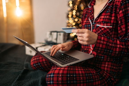 Woman shopping online in festive pajamas, holding credit card with laptop, warm Christmas lights and wrapped gifts in the background.の写真素材
