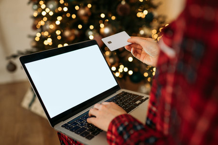 Online holiday shopping with a laptop and credit card, cozy Christmas tree lights in the background, festive ambiance.の写真素材
