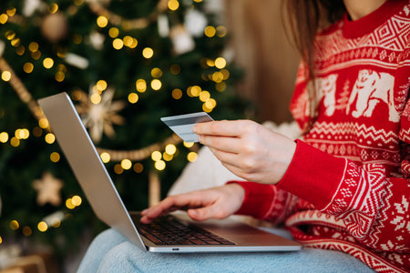 Woman in festive sweater shops online with credit card and laptop, Christmas tree lights in background.の写真素材