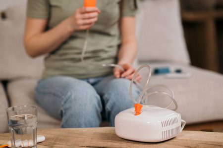 Woman using a nebulizer for respiratory treatment at home, sitting on a couch with medical equipment on the table.の写真素材