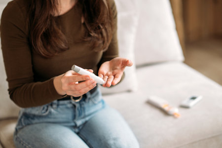 Young woman checking blood sugar level with glucometer at home, monitoring diabetes health on the couch.の写真素材